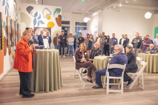 Person speaking at an event in a gallery with an attentive audience sitting and standing around tables.