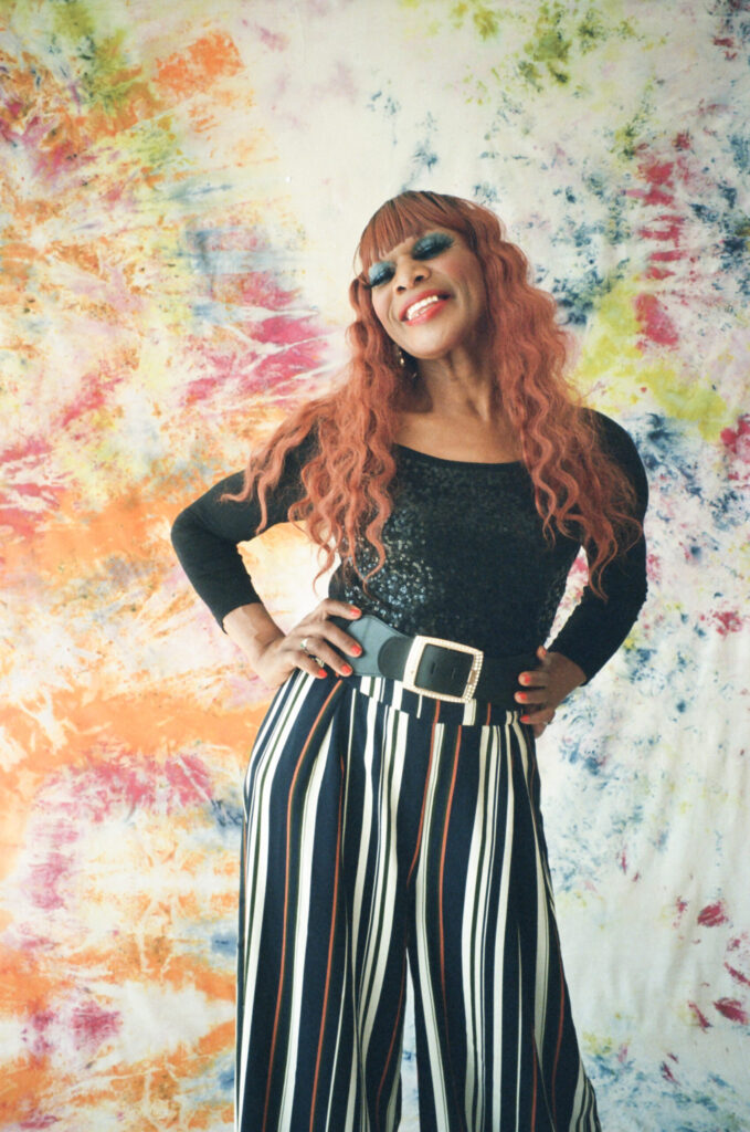 Person with long red hair, wearing a black top and striped pants, standing in front of a colorful abstract backdrop.