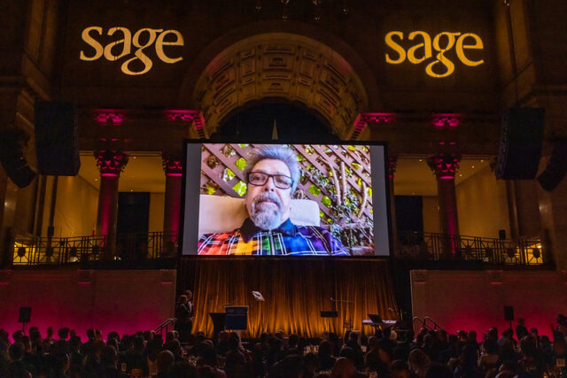 A large screen displays a bearded man during an indoor event with an audience and "sage" logos on the walls.