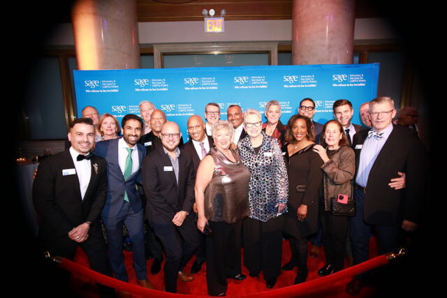 Group of people smiling and posing together in formal attire at an event with a blue backdrop and red carpet.