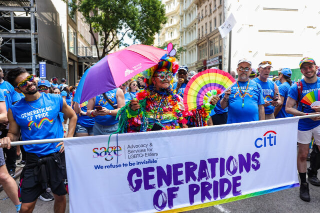 People in colorful attire holding "Generations of Pride" banner during a parade, wearing blue shirts and hats.