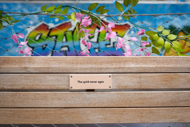Wooden bench with plaque reading "The spirit never ages," adorned with pink flowers and green leaves, colorful graffiti background.