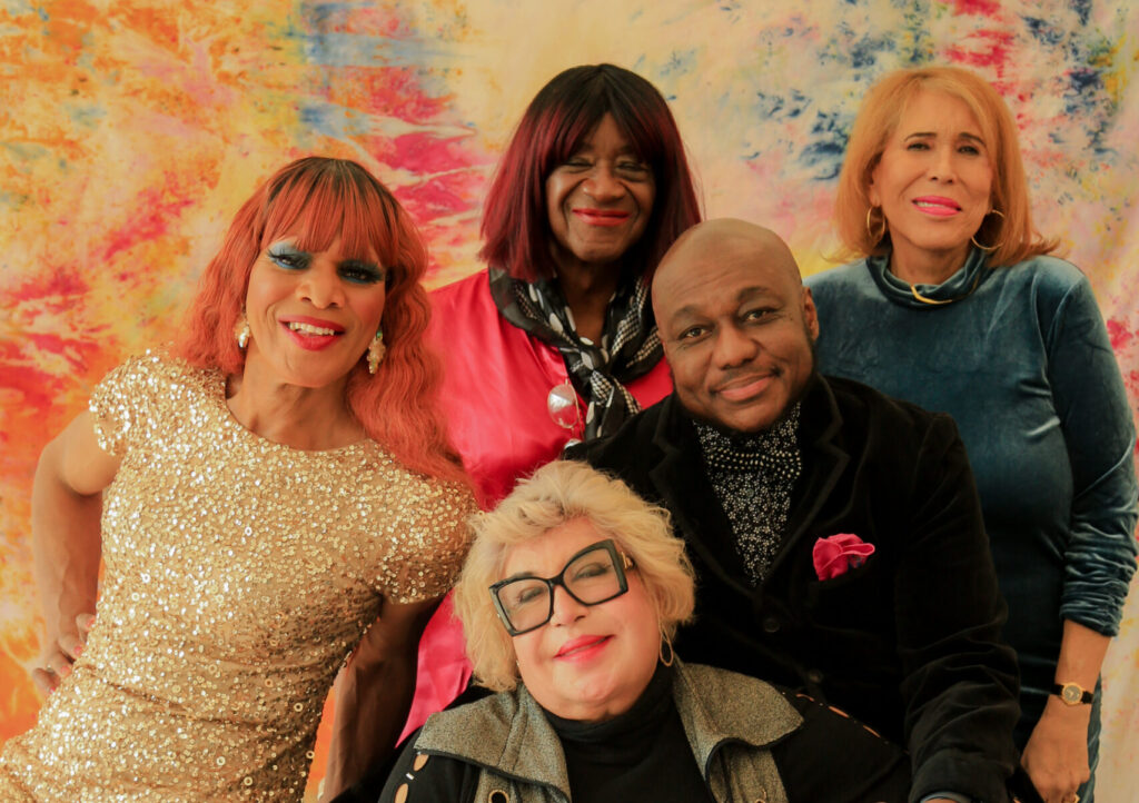 Group of five stylish individuals posing; colorful backdrop; varied expressions; seated and standing positions; vibrant and friendly atmosphere