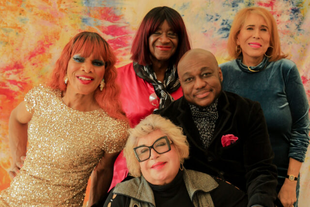 Group of five stylish individuals posing; colorful backdrop; varied expressions; seated and standing positions; vibrant and friendly atmosphere