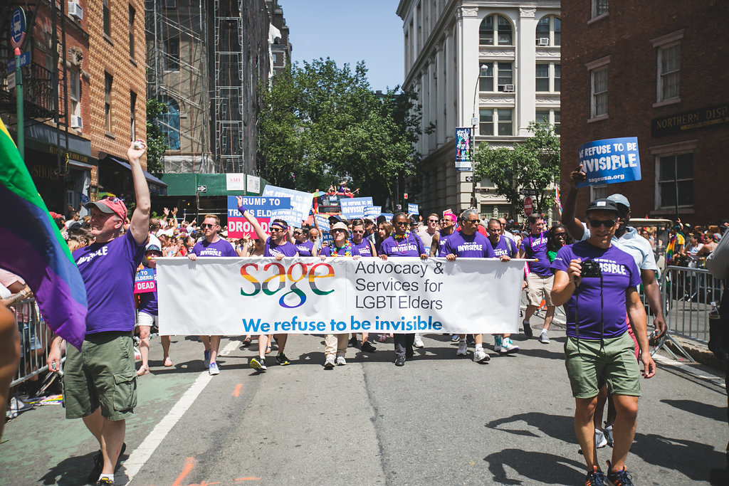 People marching in a parade holding a banner for SAGE, Advocacy & Services for LGBT Elders, with a diverse crowd around.