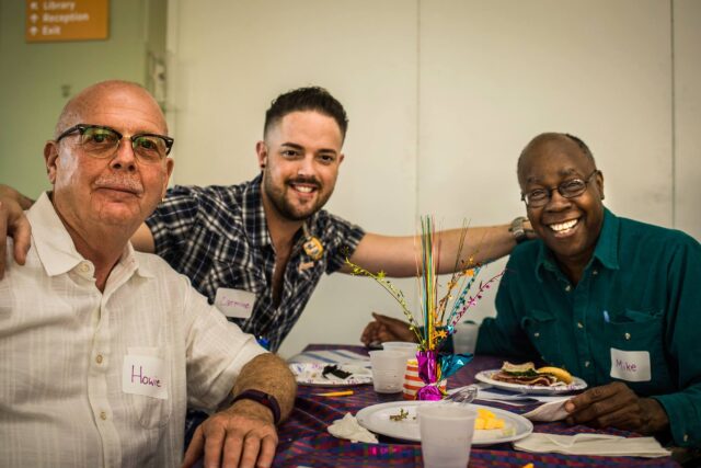 Three people smiling at a table with food and decorative centerpiece in a social setting.