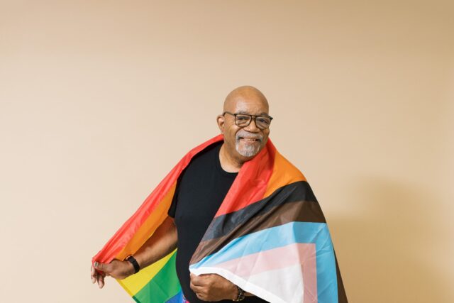 Smiling man wrapped in a colorful pride flag against a neutral background.