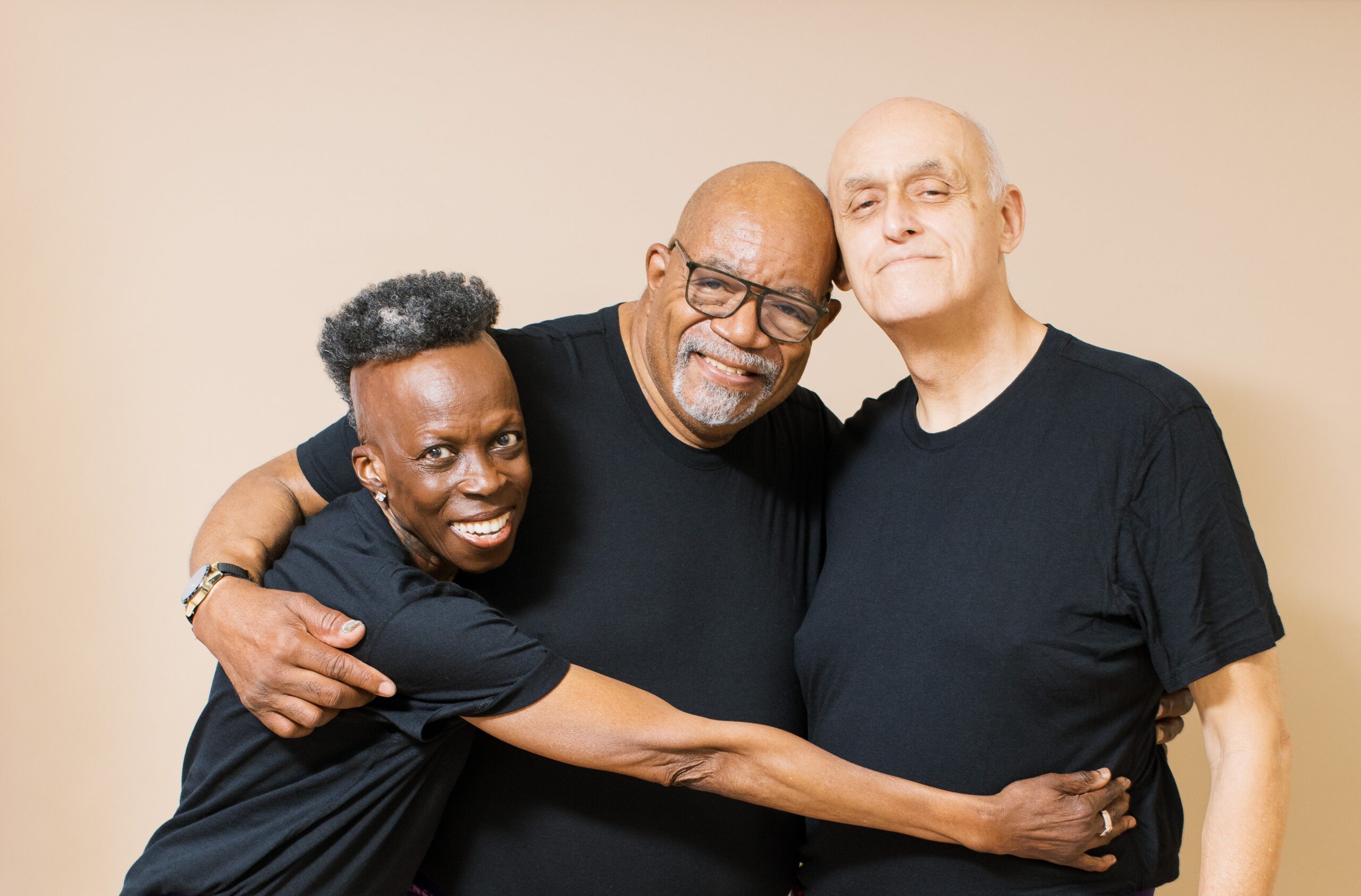 Three people wearing black shirts smiling and embracing against a beige background.